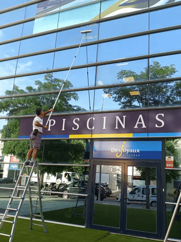 Trabajador de empresa de limpieza en Boadilla del Monte limpiando fachada acristalada de local comercial con pértiga desde escalera